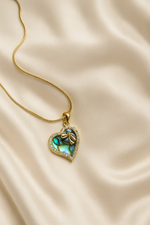 Heart-shaped pendant with green gemstone on a gold chain against a beige fabric background